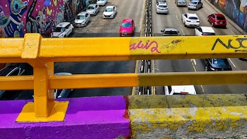 Barandilla de puente en primer plano, pintada de amarillo y morado, sobre una autopista con tráfico. A la izquierda, un muro con un mural colorido