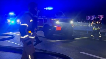 Bomberos con uniformes reflectantes caminan junto a vehículos de emergencia con luces azules en una carretera de noche