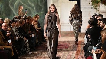 Gigi Hadid camina por una pasarela de alfombra estampada, luciendo un vestido largo de terciopelo marrón con cuello alto y hombreras de malla metálica