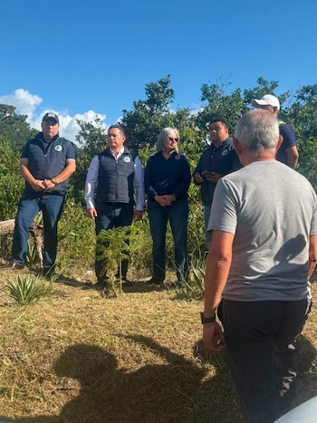 Grupo de seis personas de pie al aire libre; un hombre de espaldas en primer plano y cinco personas de frente. Un árbol pequeño está en el centro