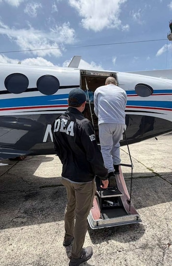 Un agente con una chaqueta con la inscripción DEA observa a un hombre de gris que sube la escalerilla de un avión en una pista al aire libre