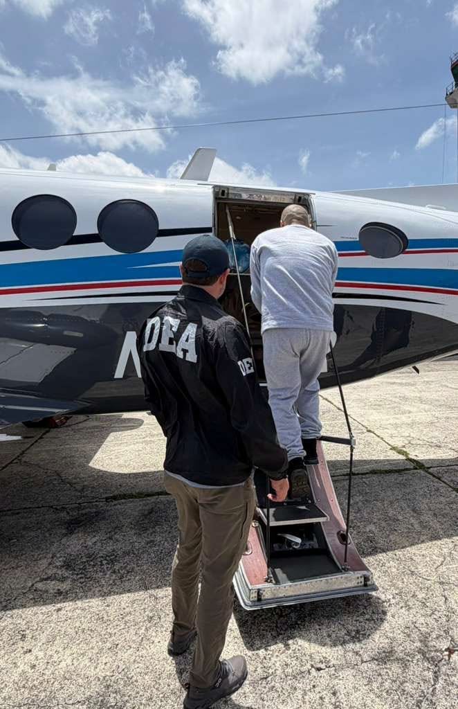 Un agente de la DEA custodia a un hombre vestido con ropa deportiva mientras este asciende por las escaleras de una aeronave en una pista de aterrizaje. (Ministerio Público de Guatemala)