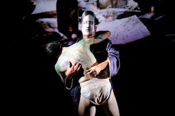 Dos artistas en el escenario oscuro; uno con máscara blanca y túnica azul sostiene a otro con pintura corporal verde/azul y calzoncillos