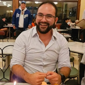 Un hombre sonriente con barba, gafas y camisa clara a cuadros está sentado a la mesa de un restaurante