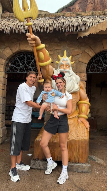 Hombre, mujer y bebé posan junto a una estatua de madera del Rey Tritón. La mujer lleva orejas de Minnie Mouse. Edificaciones de piedra de fondo