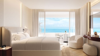 Interior de una suite de hotel con cama grande, un sillón, un mueble bajo y un balcón con mesa y sillas con vistas a un océano azul