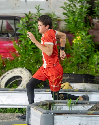 Un hombre joven con camiseta y pantalones cortos rojos, y leggings negros, corre en un circuito de obstáculos con vegetación y neumáticos al fondo