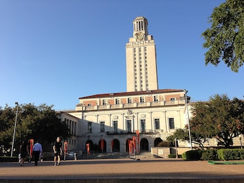 Vista exterior de la Torre y el Edificio Principal de la Universidad de Texas en Austin bajo un cielo azul, con árboles frondosos y personas caminando por la plaza