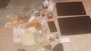 Diversos objetos, incluyendo billetes, joyas, cosméticos, dos laptops negras, un smartphone y cigarrillos, desparramados sobre el suelo