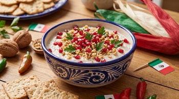 Tazón azul y blanco con crema decorada con granada, nueces y perejil, rodeado de galletas, chiles y banderas mexicanas pequeñas.