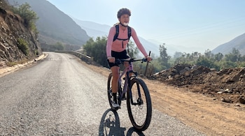 Anna Carina Copello en bicicleta por una carretera montañosa. Viste un maillot rosa, pantaloneta negra y casco, con una curita en la mejilla.