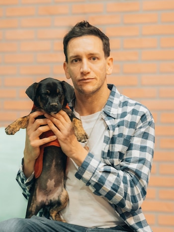 Hombre, camisa a cuadros, camiseta blanca, collar, perro cachorro de raza Dachshund, camisa roja, pared de ladrillos