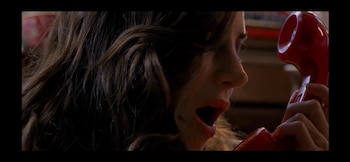 Una mujer de cabello castaño desordenado con la boca abierta sostiene un auricular de teléfono rojo, su rostro se muestra de perfil