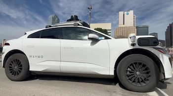 Vista lateral de un coche blanco Waymo con múltiples sensores en el techo, estacionado en una calle de la ciudad con edificios al fondo bajo un cielo azul parcial