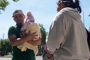 Un hombre con gorra y camiseta verde sostiene a un bebé envuelto en una manta amarilla y gorro rosa. A la derecha, una persona de espaldas. Cielo azul