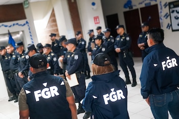 Tres personas con chaquetas oscuras que llevan la sigla FGR están frente a una fila de policías en el interior de un inmueble
