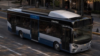 Un autobús azul y plateado con la marca Ugarte circula por una calle asfaltada junto a una acera