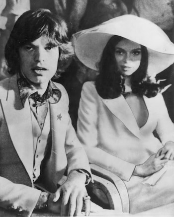 Fotografía en blanco y negro de Mick Jagger y Bianca Jagger sentados. Ella lleva un sombrero de ala ancha y traje. Él viste un traje y camisa con lazo