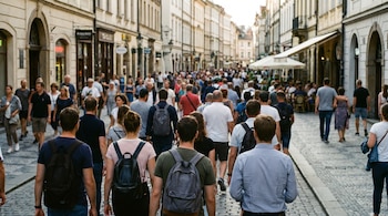 Vista trasera de una multitud de personas caminando por una calle peatonal adoquinada flanqueada por edificios. Algunas figuras al fondo están difuminadas.
