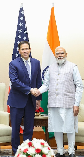 Narendra Modi, vestido de blanco con chaleco claro, y Marco Rubio, de traje azul, se dan la mano frente a las banderas de India y Estados Unidos