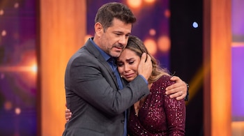 Un hombre en traje oscuro abraza a una mujer con un vestido de lentejuelas burdeos. Ella llora con la cabeza apoyada en su hombro. Fondo de estudio televisivo.