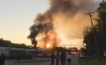 Cielo anaranjado con denso humo negro elevándose. Edificios bajos, personas observando, postes de luz, cables eléctricos y luces de vehículos de emergencia visibles