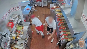 Vista aérea de dos personas con rostro pixelado agachadas en la cocina de un restaurante In-N-Out con freidoras y mostradores de preparación