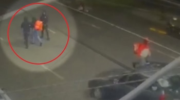 Tres personas caminan por una calle de noche, una de ellas con mochila naranja dentro de un círculo rojo, cerca de un automóvil.