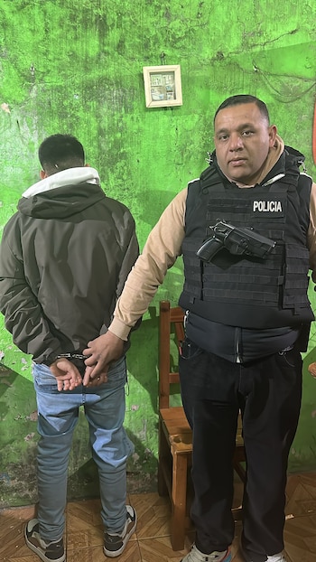Un oficial de policía con chaleco táctico y arma enfundada está junto a un hombre esposado de espaldas, contra una pared verde. Hay una silla de madera