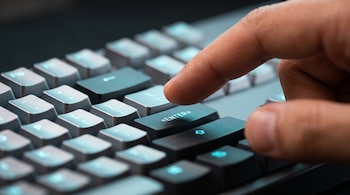 Primer plano de un dedo índice situado sobre la tecla Enter iluminada en azul de un teclado gris.