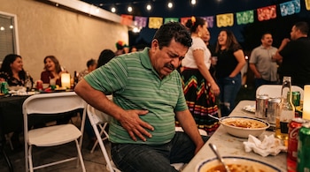 Un hombre con camisa verde a rayas se toca el abdomen junto a una mesa con comida y bebidas en un convivio nocturno decorado con guirnaldas.