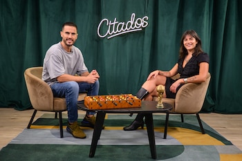 Un hombre y una mujer sentados en sillas frente a una mesa con un futbolín y un trofeo dorado. Al fondo, una cortina verde con un letrero de neón 'Citados'