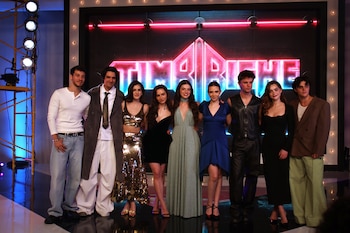 Nueve personas vestidas de fiesta posan en un escenario frente a un letrero de neón rojo con el nombre Timbiriche