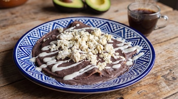 Enfrijoladas con salsa de frijol negro, queso y crema en un plato decorado, junto a una taza de café y un aguacate cortado a la mitad.