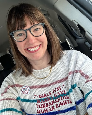 Mujer con cabello castaño, lentes, collar y suéter blanco con rayas, bordado con un texto y un distintivo de voto