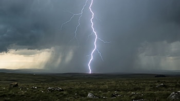 Cielo cubierto de nubes oscuras de tormenta sobre un campo de pastizales con piedras, un rayo vertical impactando el suelo, lluvia distante y horizonte pálido.