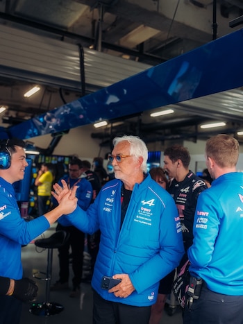 Primer plano de dos hombres chocando las manos, uno con chaqueta azul y gafas, el otro con auriculares. Personas y equipamiento de garaje en el fondo