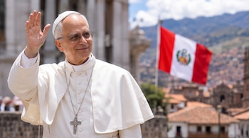 Hombre con sotana blanca, gafas y cruz pectoral que levanta la mano derecha ante una bandera de Perú al aire libre.