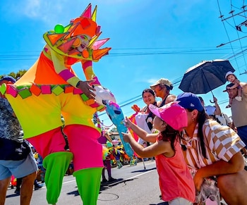 Artista en zancos con disfraz de colores neón sostiene objeto azul y se inclina hacia una niña. Adultos y público observan