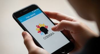 Mano sostiene un teléfono móvil con pantalla que muestra el texto 'Hate speech' y el logo de CODISRA, un círculo con figuras humanas y una cabeza.