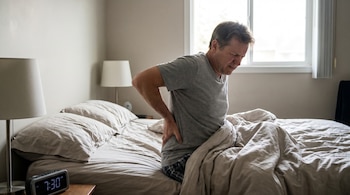 Um homem de meia-idade com uma camiseta marrom está sentado na cama, segurando a parte inferior das costas com uma expressão de dor. O despertador na mesa marca 7h30.