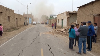 Calle de Ica con asfalto agrietado, edificios de adobe dañados y escombros. Varias personas observan los daños. Vehículos de emergencia y polvo a lo lejos.
