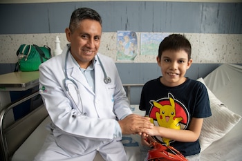 Un médico con bata blanca y estetoscopio sostiene la mano de un niño sonriente sentado en una cama de hospital