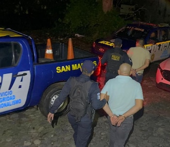 Un hombre esposado es escoltado por un policía junto a una camioneta oscura. Al fondo, un SUV rojo y un coche gris en la oscuridad de la noche