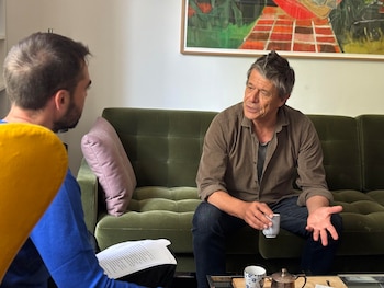 Emmanuel Carrère, de mediana edad y cabello entrecano, sentado en un sofá verde sosteniendo una taza, frente a un entrevistador joven con papeles, siendo grabado por un smartphone