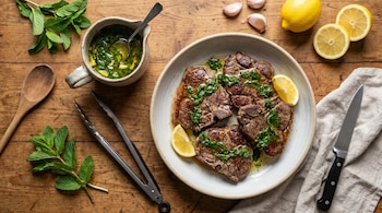 Vista cenital de bifes de cordero en un plato con rodajas de limón, salsa de menta en una jarra, ramitas de menta, limones y dientes de ajo sobre una mesa de madera.