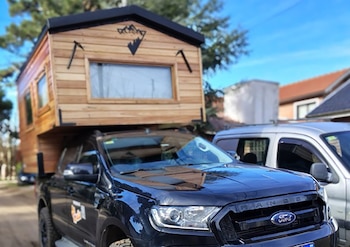 Camioneta Ford Ranger negra con módulo camper de madera y techo oscuro, ventanas laterales, logo de montaña. Otros vehículos y árboles de fondo