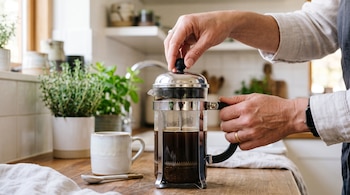 Dos manos sostienen y bajan el émbolo de una cafetera de vidrio sobre una encimera de madera, junto a una taza blanca y plantas.