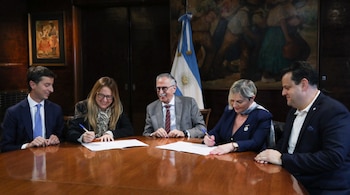 Cinco personas sentadas alrededor de una mesa de madera. Dos mujeres y un hombre firman documentos. Bandera de Argentina y dos cuadros en la pared trasera