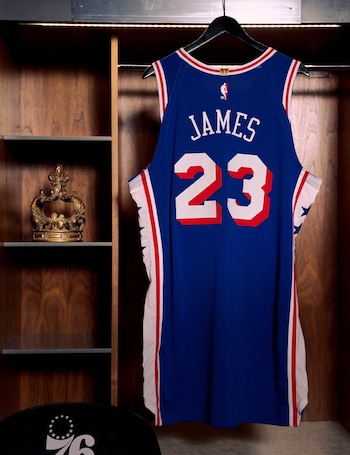 Una camiseta de baloncesto azul con el nombre James y el número 23 cuelga en un vestuario de madera, con una corona dorada en un estante y un logo 76